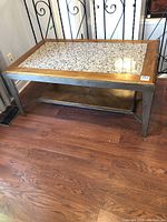 Full view of rectangular coffee table with wood frame, metal legs, stone inlay top, and wood lower shelf on hardwood floor.