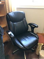 Black leather desk chair positioned near a window, showing front angle and padded armrest with white contrast stitching.