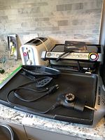 Lot on counter showing George Foreman electric griddle with attached dial, T-fal OptiGrill grill with buttons and indicator lights, black spatula, and white toaster in background.