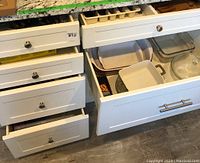 White kitchen drawers open showing ceramic and glass baking dishes including rectangular and round casserole dishes and food storage containers with lids.