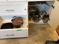 Open kitchen cabinet showing items inside: cast iron skillet, Gotham frying pan, metal cooking pots with lids, metal grater, stainless steel colander, metal bowl, and more items.