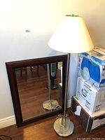 Full view of floor lamp with white shade and dark framed wall mirror leaning against the wall next to some boxed items.