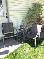 Two patio chairs placed outside on cement with a wooden plant stool between them near a house with vinyl siding