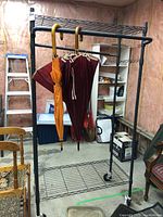 Full view of black metal rolling clothes rack standing empty aside from two umbrellas hanging from the rod, showing the rack's wire shelf bottom and upper shelf.