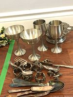 Four pewter goblets in different styles, six decorative napkin rings, ornate silverware, and cupcake candle holders on a wooden surface.
