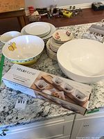 Overview of assorted serving bowls, plates with rose and gold border, and boxed snack tray set on granite counter