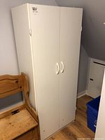 Full view of white cabinet with two closed doors and white handles, positioned beside a wooden chair.