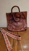 Full view of large brown leather bag with two handles and embroidered strap laid in front on wooden surface.