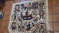 Full view of the embroidered textile showing multiple Mayan-themed figures and symbols embroidered in multicolor threads on a cream fabric base, with a zig-zag border pattern.