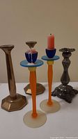 Photo showing the pair of colorful tall art glass candlesticks in front with one candle and the single heavy ornate brass candlesticks behind them