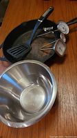 Photo showing large cast iron frying pan with black spatula inside, stainless steel bowl nearby on wooden table.