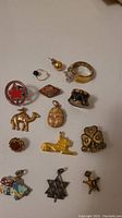 Photo showing a collection of 14 assorted charms and pins, including a silver maple leaf charm with red enamel, gold camels, scarab, lion charms, a drum charm, butterfly ring, and other decorative charms and pins.