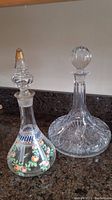 Side view showing hand-painted floral decanter with gold-accented stopper standing next to pressed glass starburst decanter with round glass stopper.