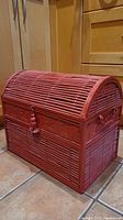 Side angled view of red glossy painted metal and bamboo hinged trunk on tiled floor in front of wooden cabinet.