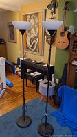 Pair of tall black metal floor lamps with frosted white globe shades on round bases, situated in a living room with piano, guitar, and artwork in background.