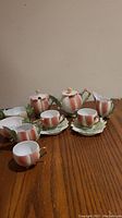 Full view of the porcelain tea set on wooden table showing teapot, sugar bowl, creamer, teacups and saucers with intricate gilt handles and leaf motifs.