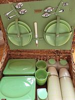 Top view of inside lid showing four metal spoons, one knife, two olive green plates secured with straps. Below the lid, green plastic canisters, cups, and two thermoses with beige bodies and green lids.
