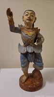 Front view of the painted wooden Thai statue showing detailed facial features, clothing colors, and raised hand gesture.