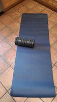 Blue yoga mat laid out on tile floor with black PURAthlete3 foam roller placed on it.