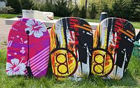 Three body boards: one pink Surfgear board with floral design and safety strap, two Banzan boards with black and orange graphics, one missing safety strap.