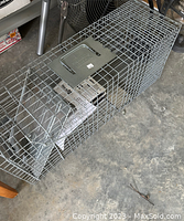 Photo of the Havahart cage trap showing wire mesh and handle from top view, placed on a concrete floor with some other items around.