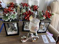 Six black and gold holiday lanterns with snow-covered winter village scenes inside, each topped with artificial pine sprigs, red bows, and berries, grouped on a floor with a chair in the background.