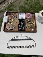 Full view of jewelry displayed on metallic tray on a table, containing various earrings, bracelets, pins, and necklaces along with a black plastic hair comb and a white container in background.