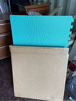 Green foam floor mat partially out of a worn cardboard box, showing interlocking puzzle edges and textured surface.