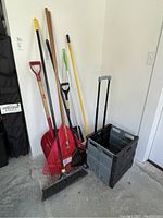 Various garden and cleaning tools including brooms, shovels, rake, and foldable rolling cart with handle, arranged against a white wall on garage floor.