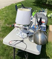 Photo showing two lamps on a table outdoors on grass. One lamp has a white shade and horse-shaped base, the other is metal with adjustable arm.