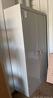 Tall gray metal cabinet with two doors and lock, seen from a left-side view, standing next to a wall.