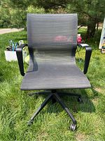 Front view of the black mesh office chair on grass, showing seat and backrest with armrests and five caster wheel base.