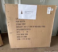 Photo of cardboard box labeled LB International with printed label showing a 6ft red and gold pop up tree image and details.