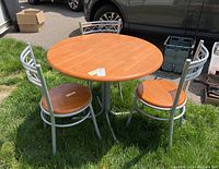 View of round table with wood top and metal base, with three matching chairs arranged around it on grass outside.