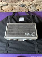 Closed view of black and silver aluminum carrying case with Jackpot logo on top with handle and latches.