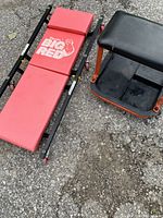 Red padded folding under car creeper and black mechanic stool placed on asphalt surface.