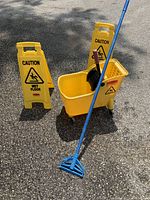 Wide view of the Mop Bucket with wringer, blue mop handle leaning against it, and two yellow caution signs in the background on paved ground.