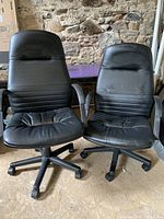 Two black desk chairs side by side on a rough floor, showing fronts with visible wear and creasing.