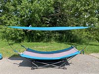 Blue and gray portable hammock assembled outdoors with attached blue sun shade and black metal frame on grass with trees in background.