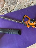 Orange and black WORX hedger trimmer with blade and plastic cover on purple table, stone wall background.