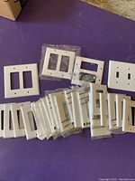 Several white outlet and switch cover plates displayed on a purple surface. Some are loose while others remain factory-sealed in plastic packaging.