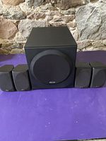 Full set of Polk Audio speakers and subwoofer displayed on a purple surface against a stone wall.