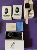 Four smartwatches shown: three boxed Smart Watch Your Sports Partner packages and one beige smartwatch outside the box on purple fabric.