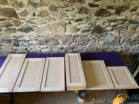 15 solid maple door faces, varying sizes, light wood grain with panel design, laid out on a table indoors against stone wall.