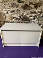 White wooden rectangular storage chest with a closed hinged lid against a stone wall background, placed on a purple surface.