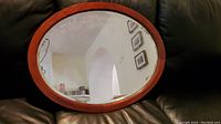 Front top angled view of oval wooden framed mirror reflecting wall and pictures