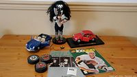 Overview photo showing KISS statue, blue Porsche model car, red Volkswagen Beetle model, two hockey pucks, and two magazines on wooden surface