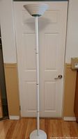 Full white metal floor lamp standing against a door, showing the overall height and slender pole design.