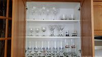 Glassware arranged on three shelves inside a wooden cabinet including tall and short glasses, some with decorative details.