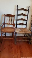 Photo of two wooden ladder back chairs with woven rattan seats side by side on hardwood floor against a wall.